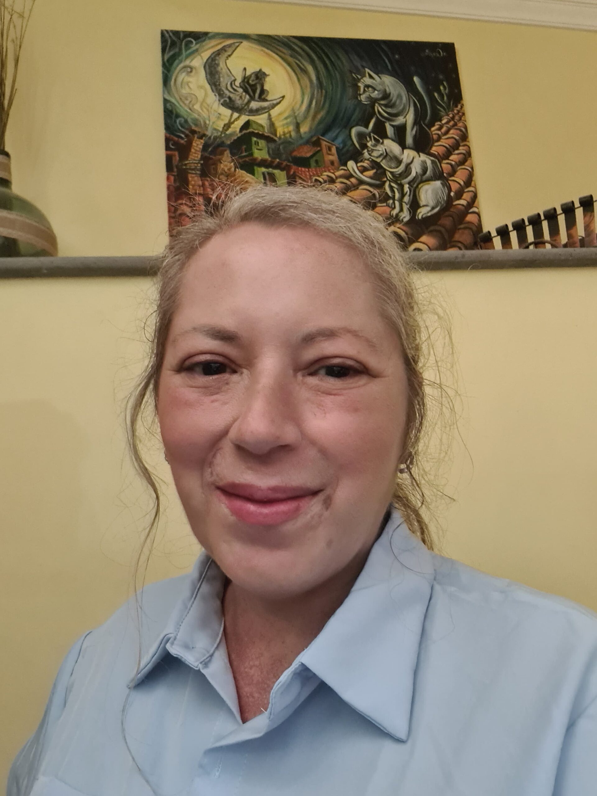 Mujer sonriente en primer plano, vistiendo una camisa de color azul, con un fondo de pared amarilla y una obra de arte colorida que representa gatos y un paisaje urbano.