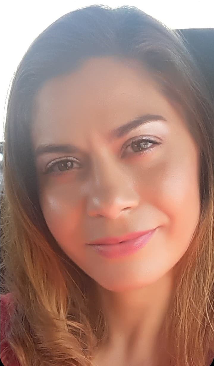 Retrato en primer plano de una mujer con cabello largo y ondulado, sonriendo suavemente. La imagen muestra un fondo difuso, destacando su rostro iluminado y natural.