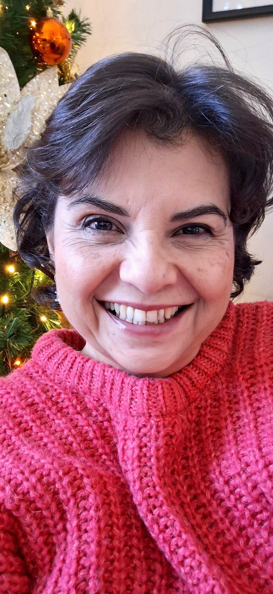 Mujer sonriendo con un suéter rosa, frente a un árbol de Navidad decorado con luces y adornos.